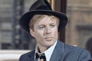 A man in a vintage suit, striped tie, and fedora hat sits indoors with a serious expression, chewing a toothpick. A blurred car and building are visible outside the window behind him.