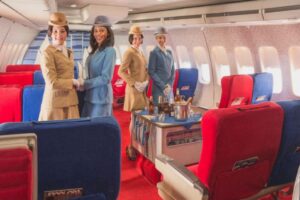Four flight attendants in vintage uniforms stand and smile inside a retro airplane cabin with red and blue seats, a service cart with drinks, and large windows along the wall.