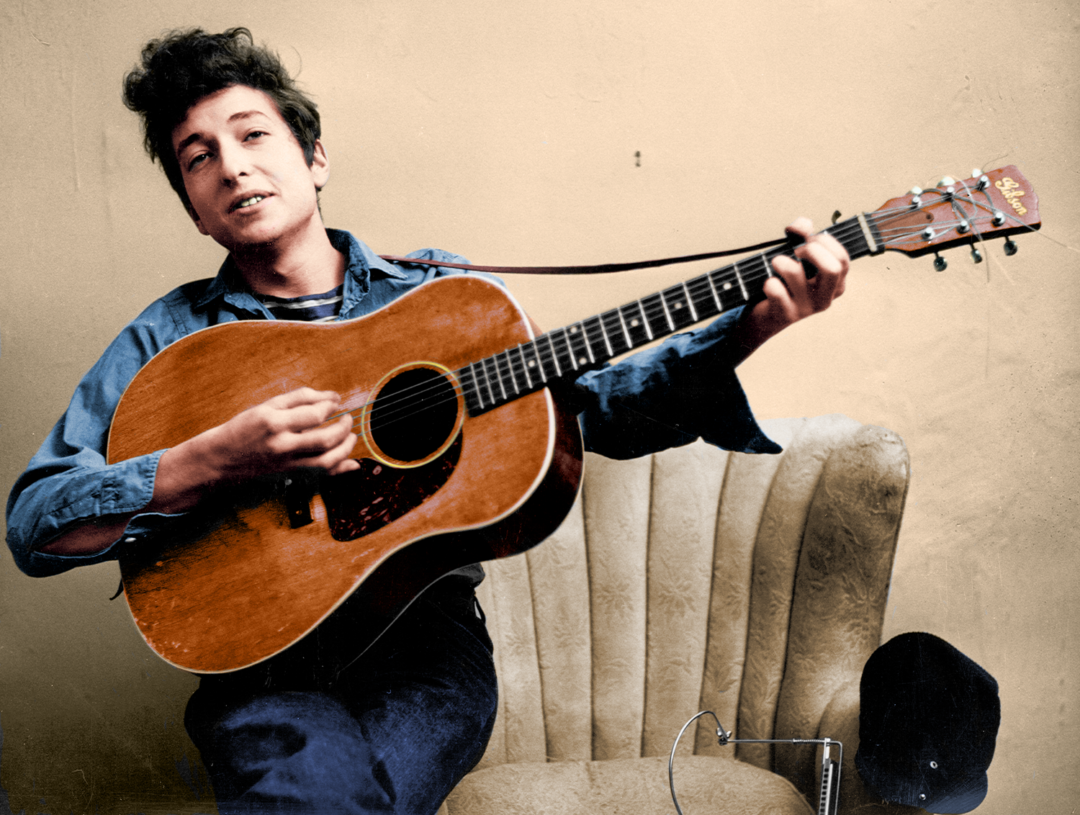 Young man with tousled hair sits on a beige chair, strumming an acoustic guitar. He wears a blue button-up shirt and dark pants. A black hat and audio recorder rest on the chair beside him. The background is plain.
