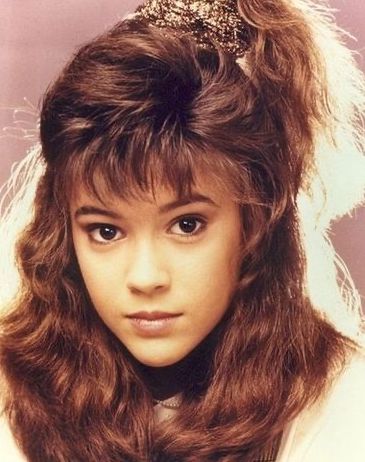 A young girl with long, wavy brown hair styled with a high side ponytail and bangs, looking directly at the camera with a neutral expression against a soft pink background.