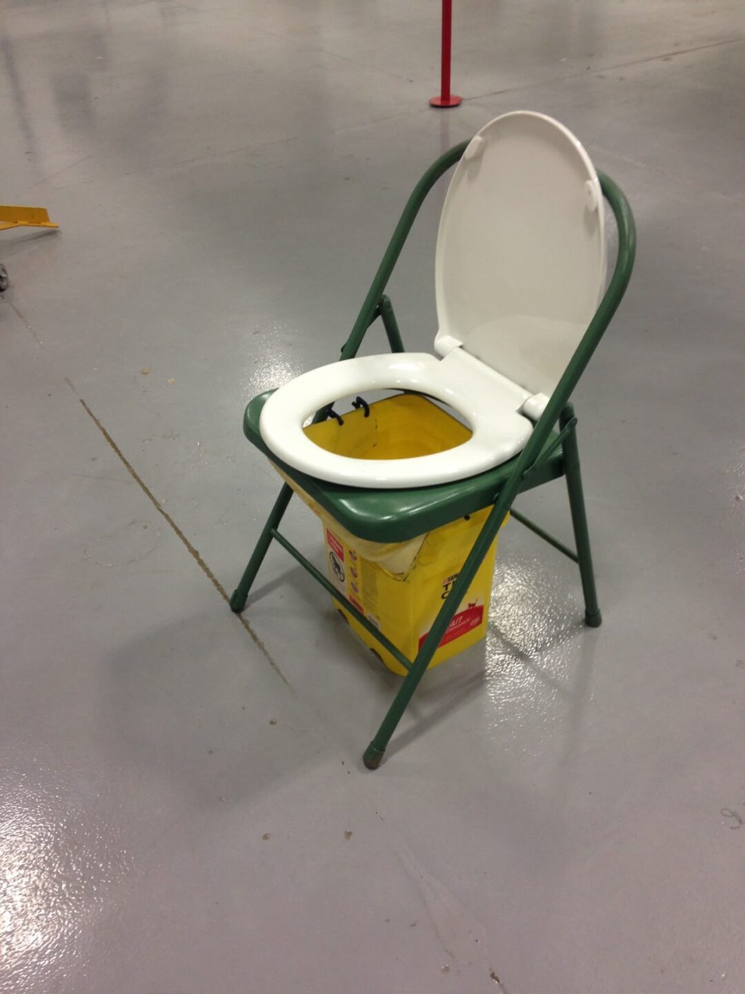 A green folding chair with a toilet seat attached, placed over a yellow mop bucket, set on a gray indoor floor.