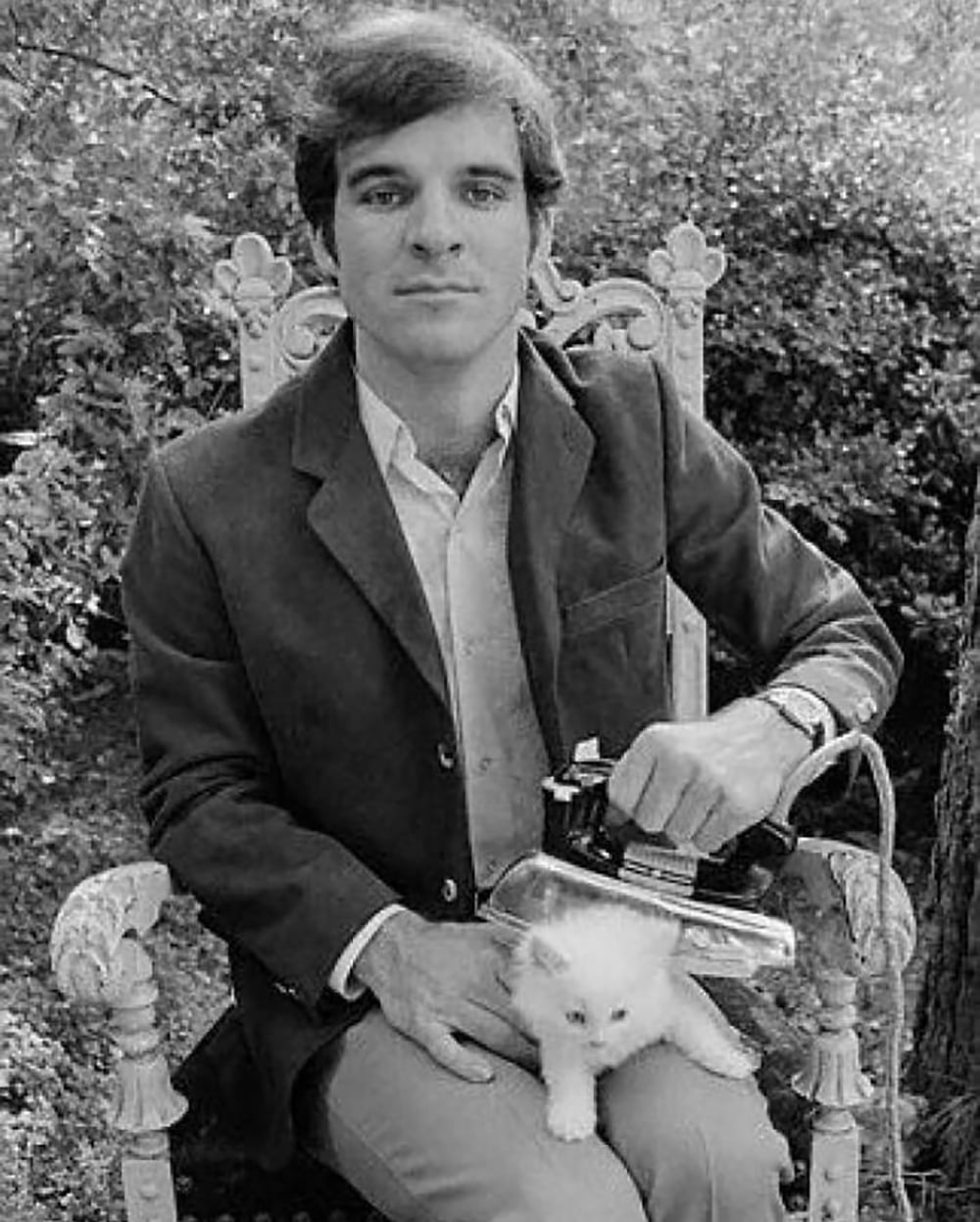 A man in a suit sits on an ornate chair outdoors, holding an iron over a white kitten on his lap. The scene is black and white and appears playful and staged. Trees and bushes are visible in the background.