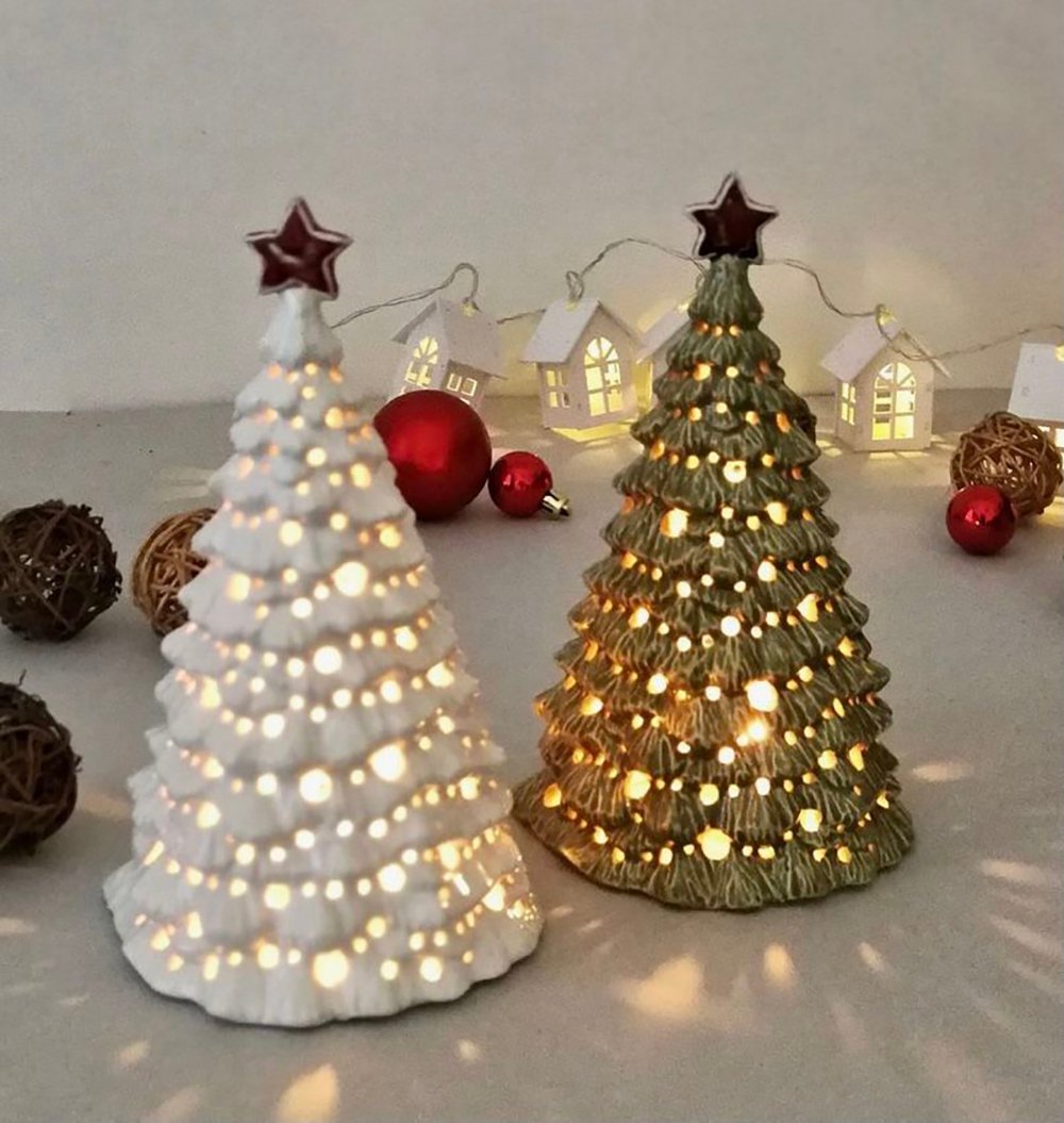 Two decorative Christmas tree ornaments, one white and one green, both with star toppers and glowing lights. In the background are string lights with small house shapes, red ornaments, and rattan balls.