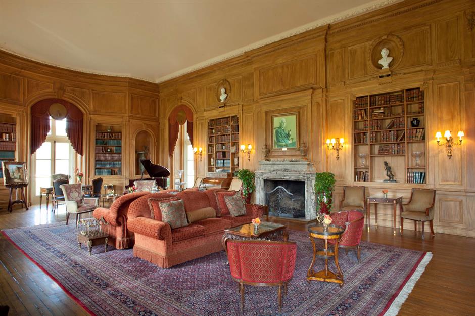 A spacious, elegant library with wood-paneled walls, built-in bookshelves, a grand piano, ornate fireplace, classic furniture, large windows, and an elaborate rug covering the wooden floor.