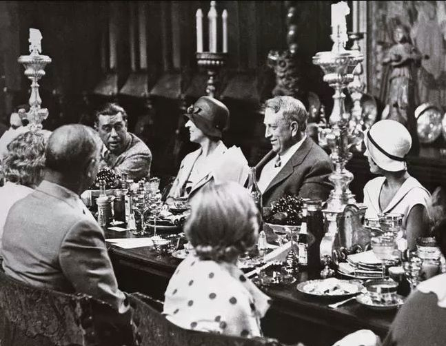A black-and-white photo of people in vintage attire seated around an ornate dining table, engaged in conversation. The table is set with candles, glassware, and silverware, and the room has an elegant, classic decor.