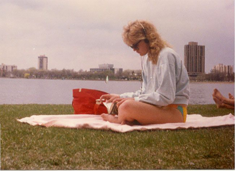 A woman in sunglasses and headphones sits on a blanket by a lake, reading a book with a red bag beside her. She is wearing a sweatshirt and swimsuit bottoms. Buildings and trees are visible across the water.