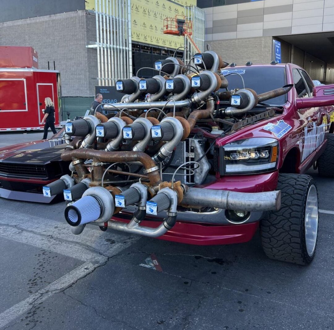 A red pickup truck with its hood removed, displaying an elaborate engine setup featuring multiple large turbochargers and air filters, parked at an outdoor event near modern buildings.