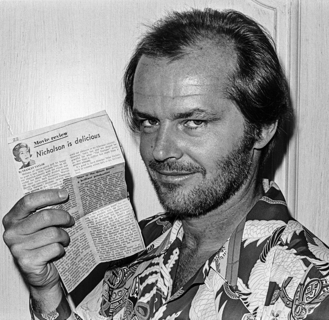 A man with a beard and receding hairline, wearing a patterned shirt, smiles and holds up a newspaper clipping with the headline “Nicholson is delicious.”