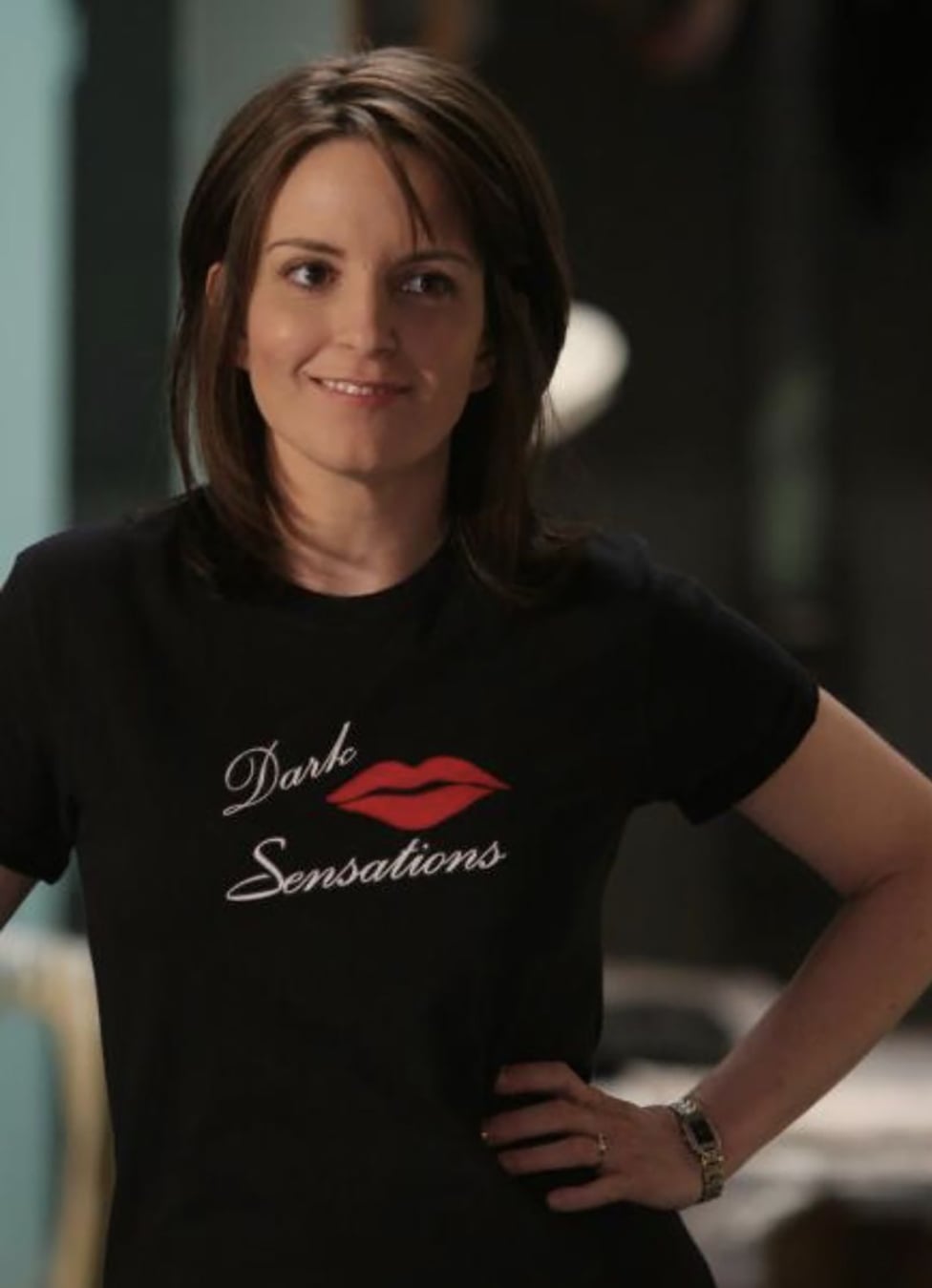 A woman with straight brown hair smiles slightly, wearing a black t-shirt with a red lips graphic and the words "Dark Sensations" written in white script. Her hands rest on her hips.