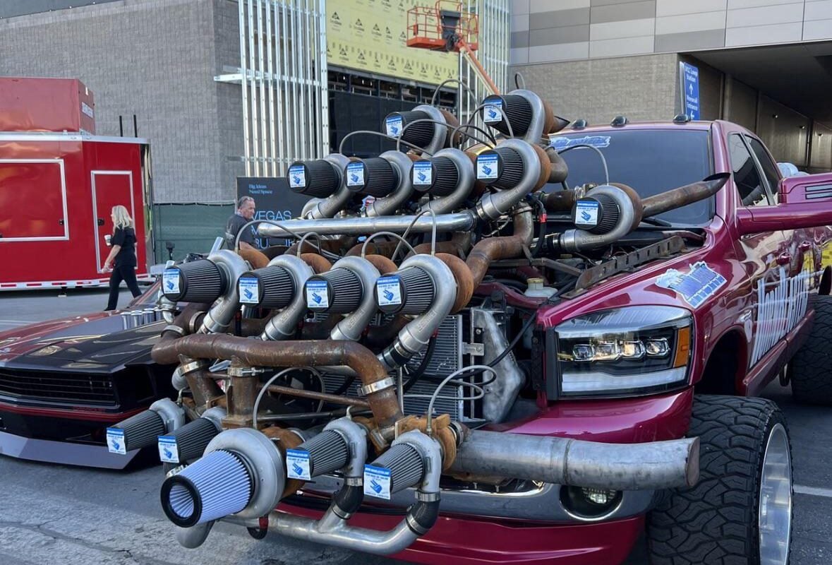 A red pickup truck with an exaggerated setup of many turbochargers mounted on its hood, parked outdoors near modern buildings. The truck's front end appears heavily modified and eye-catching.