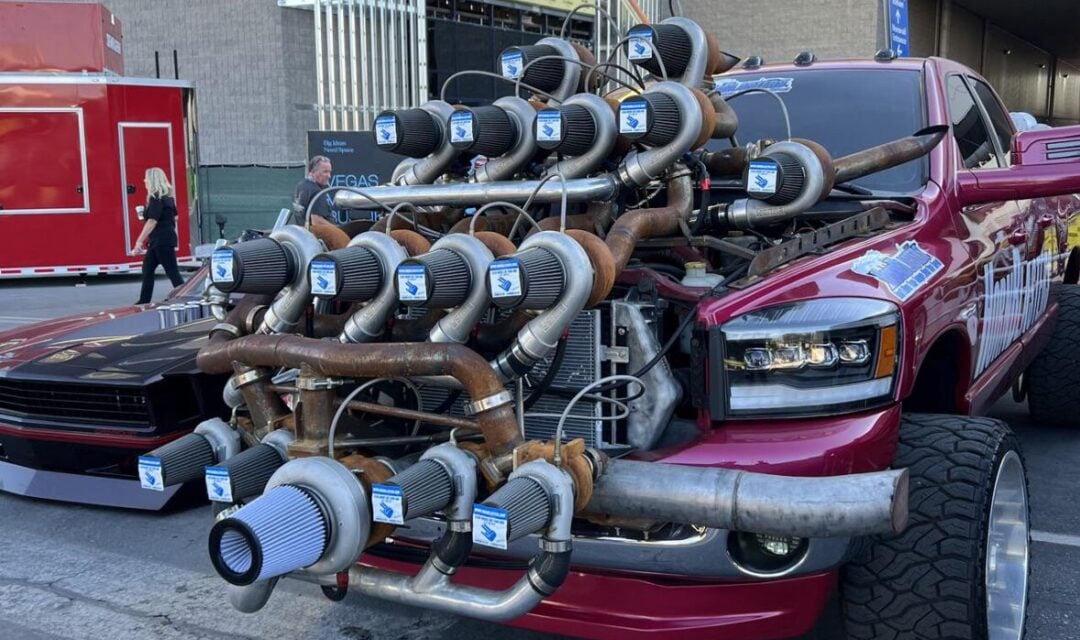 A red pickup truck with an exaggerated setup of many turbochargers mounted on its hood, parked outdoors near modern buildings. The truck's front end appears heavily modified and eye-catching.