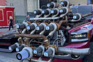 A red pickup truck with an exaggerated setup of many turbochargers mounted on its hood, parked outdoors near modern buildings. The truck's front end appears heavily modified and eye-catching.