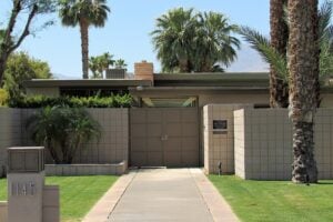 A mid-century modern house with a flat roof, beige concrete block walls, and a gated entry. Palm trees and green lawn surround the home. A mailbox with the number 1145 is visible in the foreground.