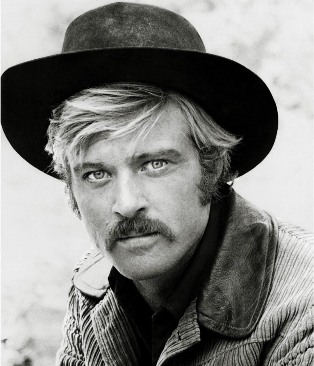 Black and white portrait of a man with light hair and a mustache, wearing a wide-brimmed black hat and textured jacket, looking directly at the camera with a serious expression.
