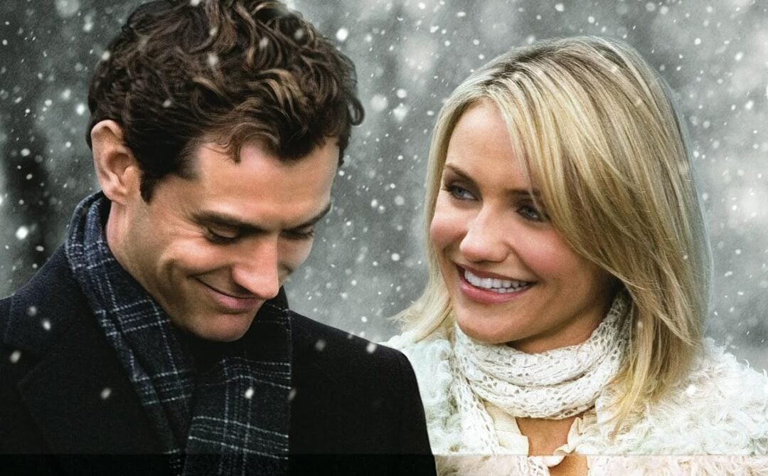 A man and a woman stand close together outside in falling snow. The man looks down and smiles while the woman looks at him, smiling warmly. Both are dressed in winter clothing, including scarves and coats.