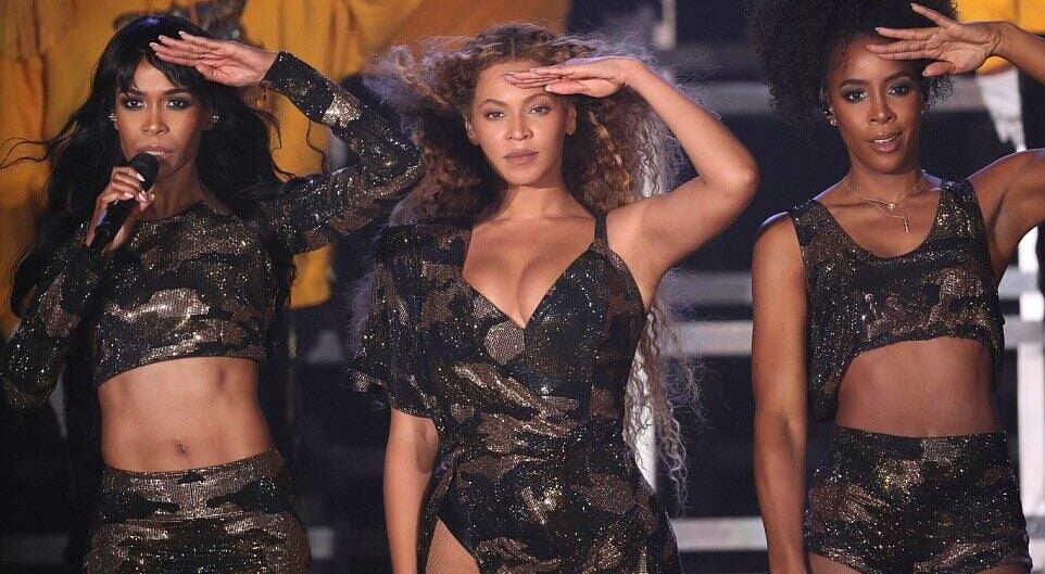A performer in an elaborate, sparkling black and gold cape, jeweled bodysuit, and ornate headdress stands confidently on stage holding a staff, with dramatic lighting and a dark background.