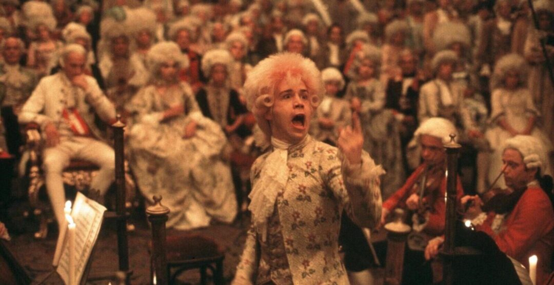 A man in an elaborate 18th-century costume and white wig passionately conducts an orchestra before a large, attentive audience dressed in period attire and powdered wigs. The scene is richly lit by candlelight.