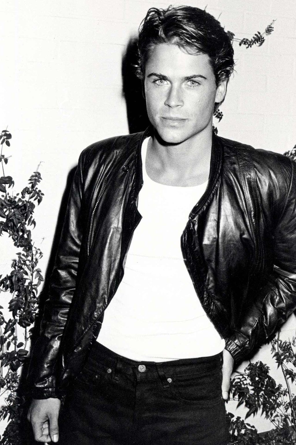 A young man with short hair wears a black leather jacket over a white t-shirt and dark pants, standing against a light-colored wall with some leafy plants on the side. The photo is in black and white.