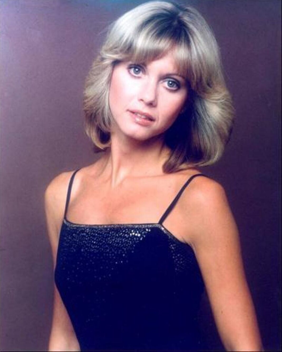 A woman with shoulder-length blonde hair and bangs wears a black spaghetti strap dress with sparkly details, looking at the camera against a plain brown background.