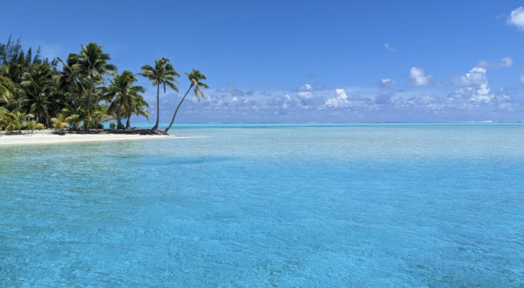 A tropical beach with clear turquoise water, white sand, and palm trees under a bright blue sky with scattered clouds. The peaceful scene suggests a remote island paradise.