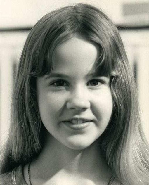 A black and white close-up photo of a young girl with long, straight hair and a slight smile, looking directly at the camera. The background appears blurred.