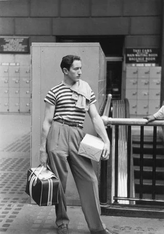 A man wearing a striped shirt and high-waisted pants stands in a train station, holding a small package and a striped bag. He has a sweater draped over his shoulder and looks to the side, with lockers and signs in the background.