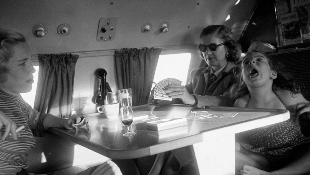 Three women and a girl sit around a table in an airplane cabin, playing cards. One woman deals cards, while the girl next to her opens her mouth wide in a dramatic expression. Sunlight streams through the window.
