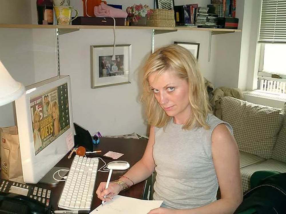 A woman with blonde hair sits at a desk, holding a pen and writing on paper. She looks up at the camera. A computer, lamp, and office supplies are on the desk, with shelves and a window in the background.