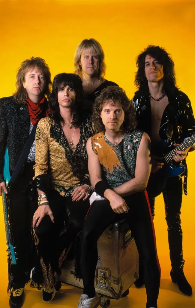 Five men pose together against a bright yellow background. Some are holding instruments, and they wear flashy, rock-style clothing, with sequins, animal prints, and open shirts. They appear to be members of a rock band.