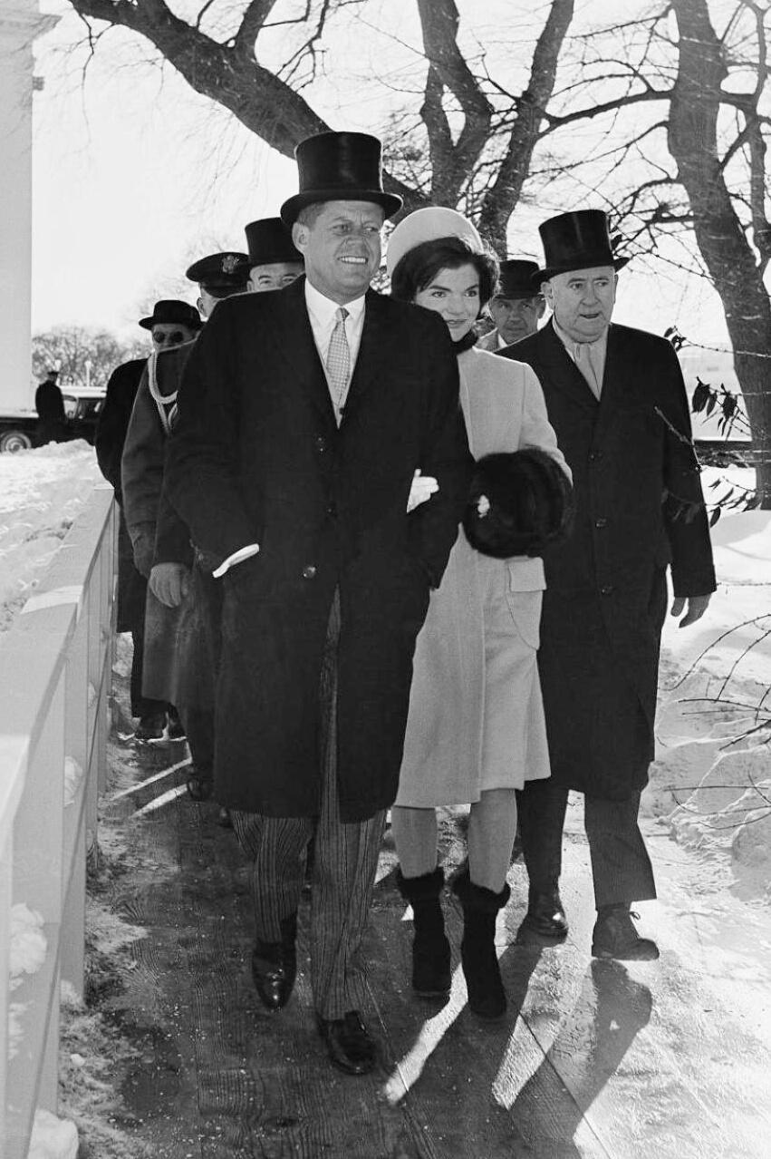 A group of people in formal winter attire, including long coats and top hats, walk outside on a snowy path. The woman in front wears a light coat and holds a fur muff. Bare trees and snow are visible in the background.