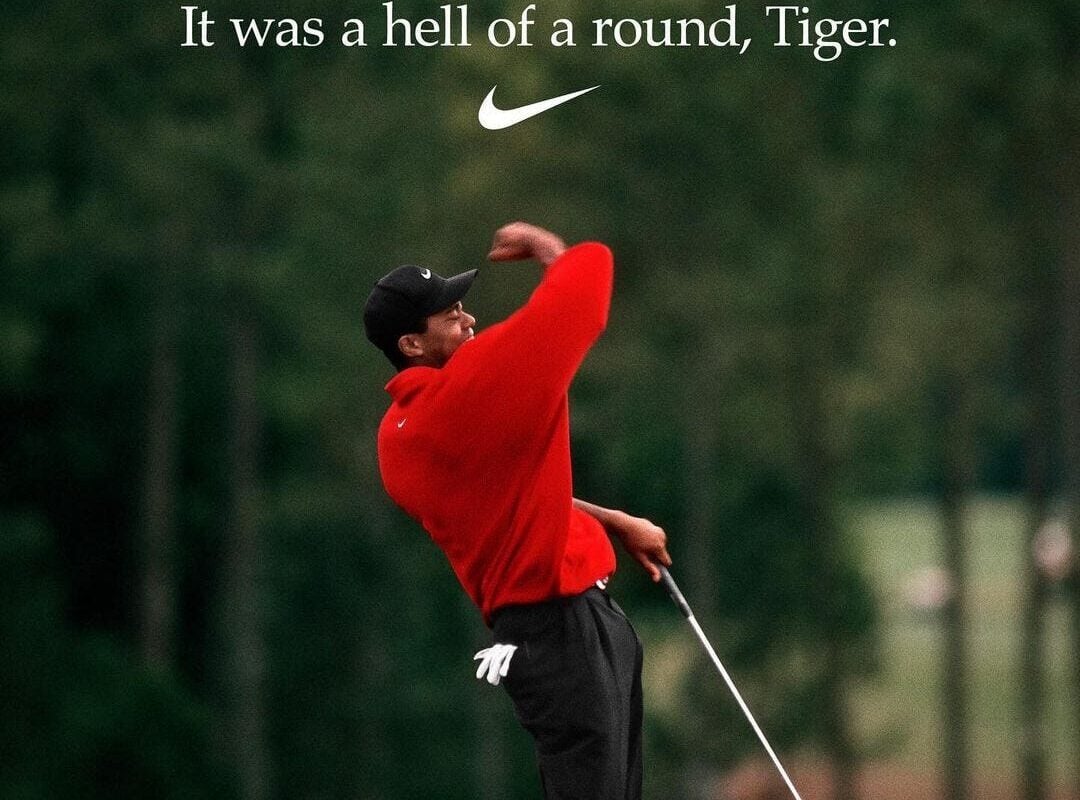 A golfer in a red shirt and black pants celebrates a successful shot on a course, fist raised. Text above reads, "It was a hell of a round, Tiger," with a Nike logo below the text. Trees and blurred spectators are in the background.