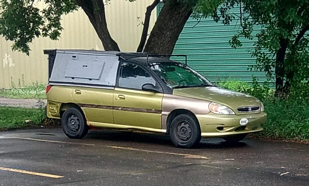 A woman with short gray hair poses confidently in a long-sleeved, shimmering gold sequined dress, raising one arm slightly above her head. The background is dark and plain.
