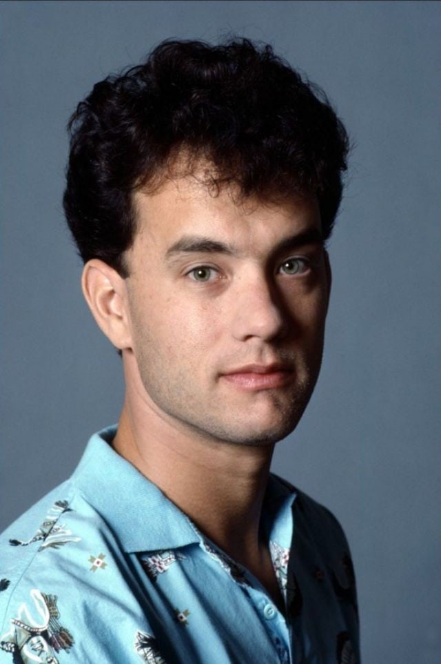 A young man with short, dark curly hair wearing a blue shirt with a subtle pattern, looking directly at the camera against a plain blue background.