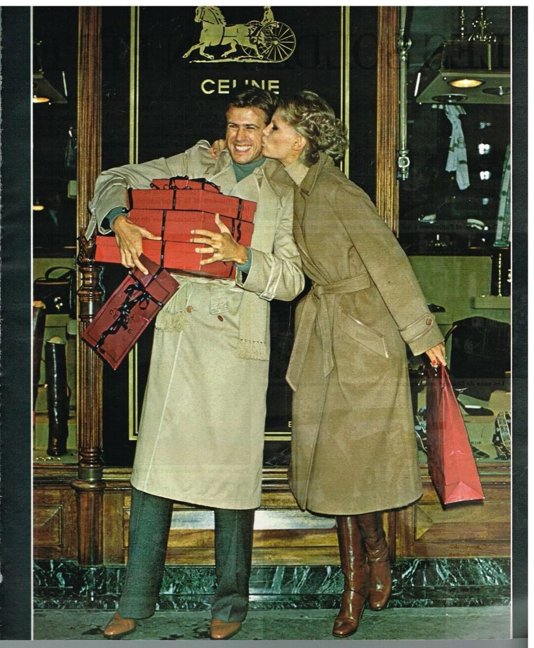 A smiling couple in tan trench coats stands outside a luxury store. The woman kisses the man on the cheek as he holds several red gift boxes. She carries a shopping bag and wears brown boots.