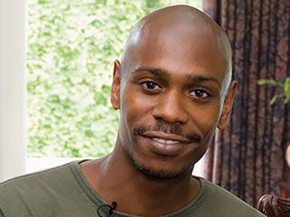 A man with a shaved head and short mustache smiles softly at the camera. He is wearing a green shirt and is indoors, with a blurred background of a window and patterned curtains.
