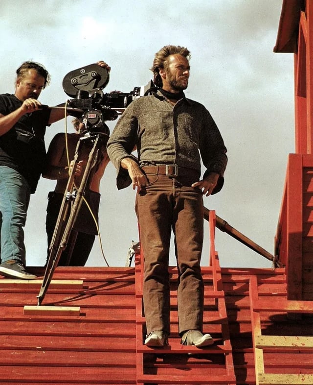 A man in casual clothes stands confidently on red wooden steps beside a camera crew filming with a large movie camera under a cloudy sky.