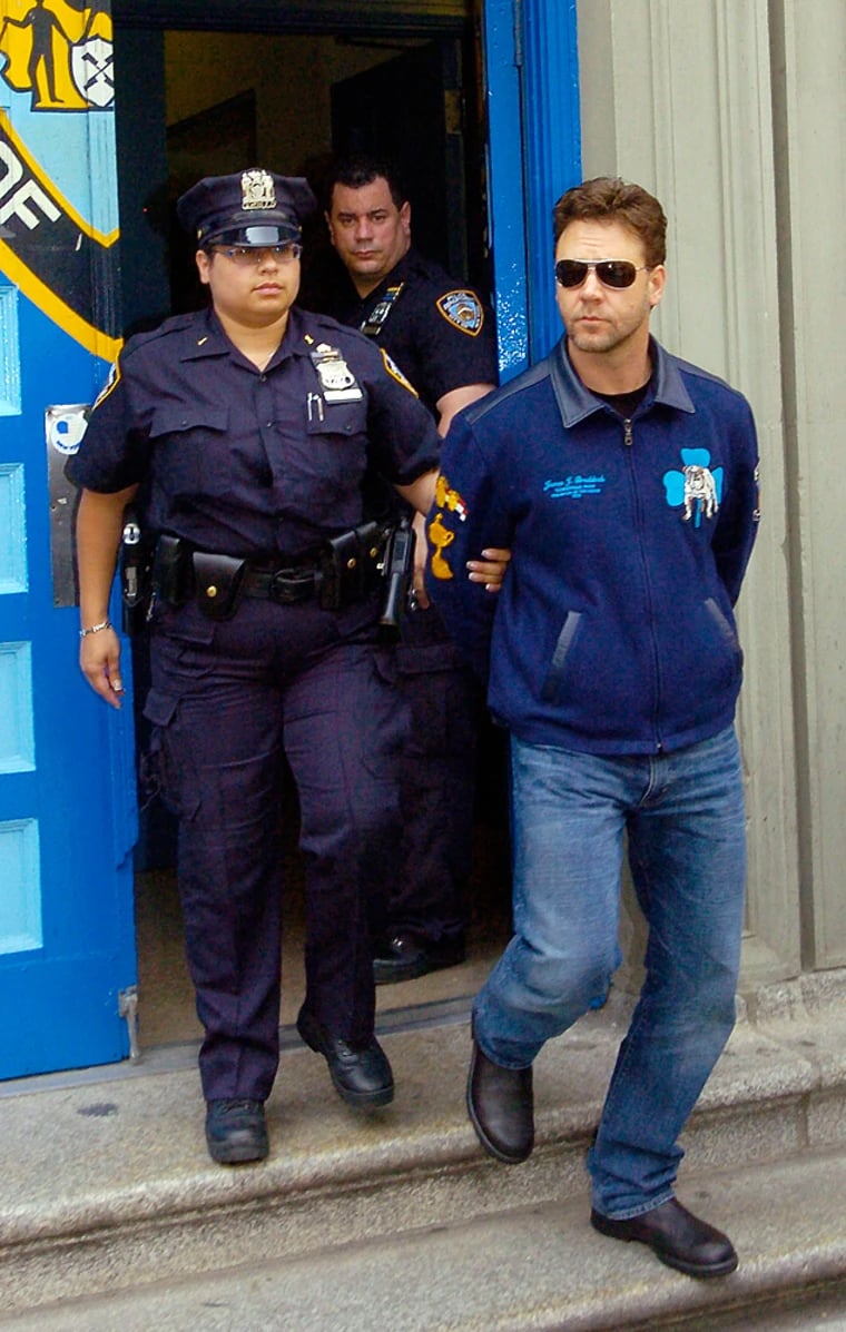 A police officer escorts a handcuffed man in sunglasses and a blue jacket out of a building, while another officer stands in the doorway behind them.