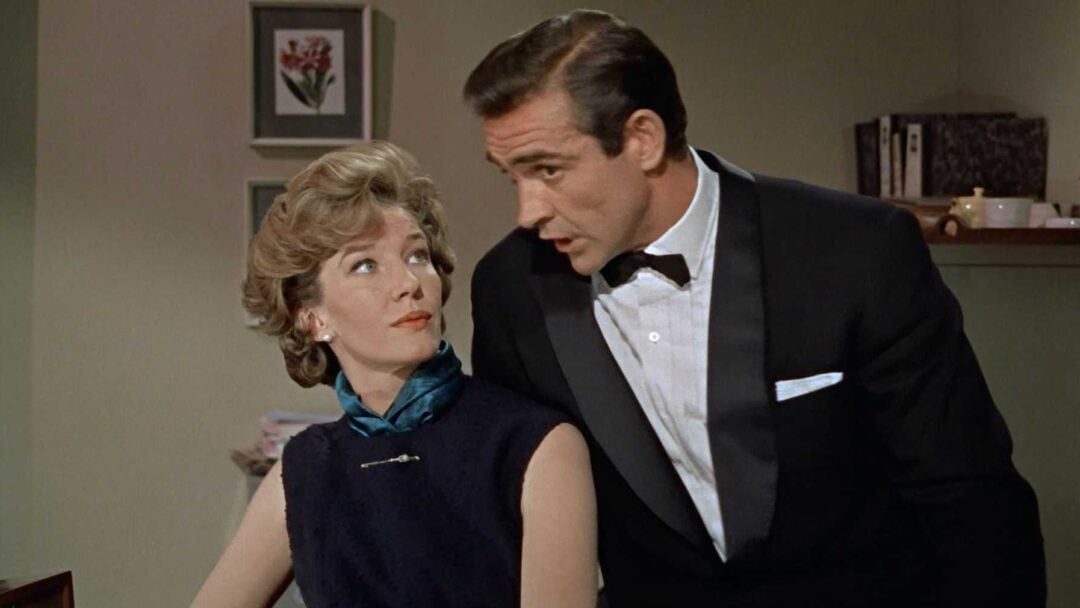 A woman with short blonde hair in a dark dress and blue scarf sits at a desk, looking up at a man in a black tuxedo and bow tie who is leaning toward her. They appear to be having a serious conversation indoors.