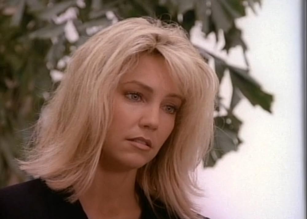 A woman with shoulder-length blonde hair and light makeup looks slightly downward with a serious expression. She is outdoors, and a leafy green plant is blurred in the background.