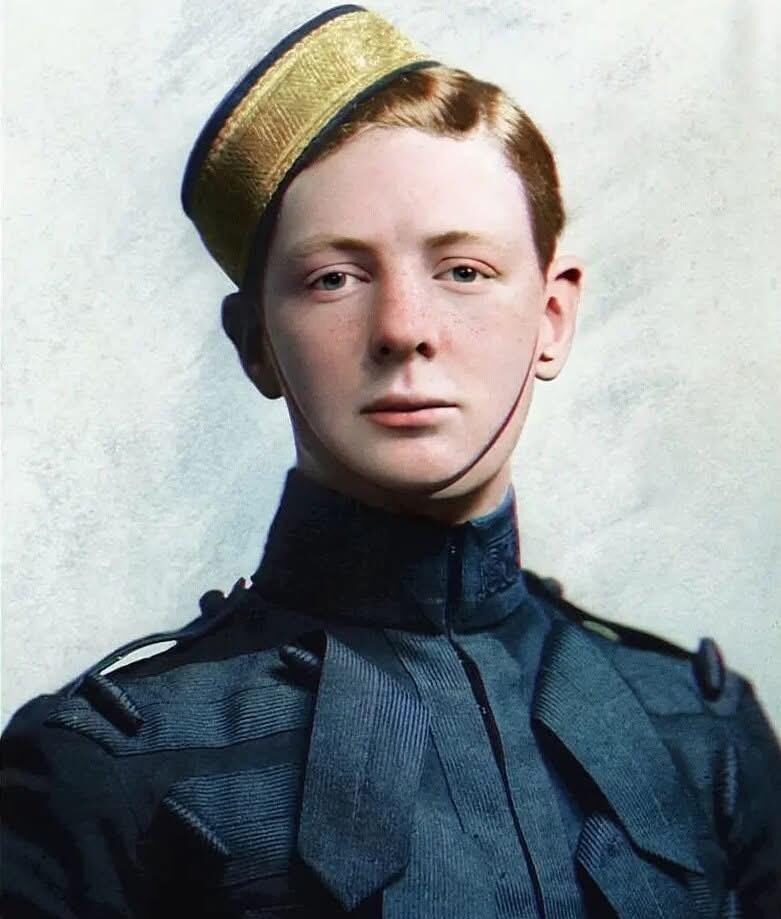 A young man in a formal military uniform with decorative braiding and a tall, cylindrical hat with a gold band poses for a portrait against a plain background.