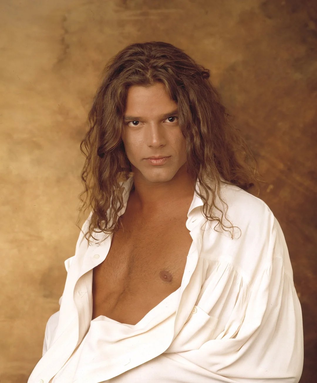 A person with long, wavy brown hair wearing a loose white shirt partially open at the chest poses against a warm brown textured background, looking directly at the camera.