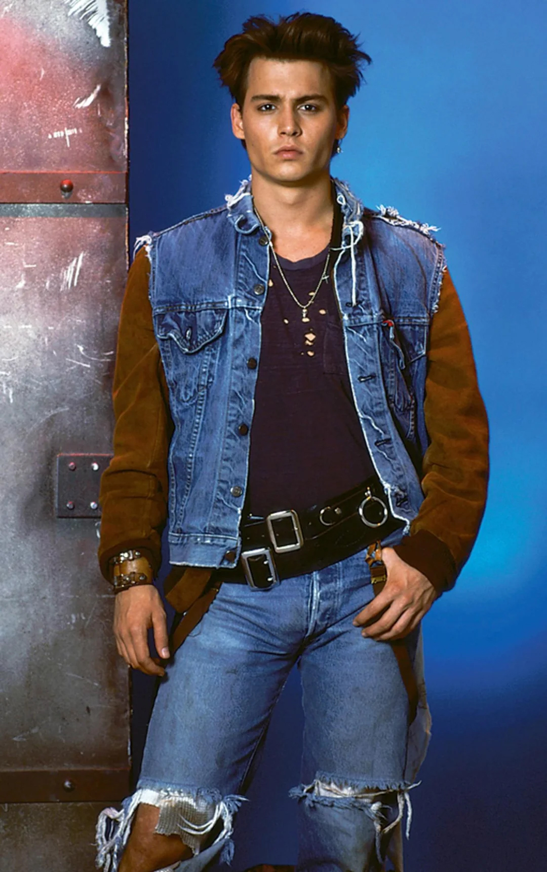 A young man with dark hair stands against a blue background, wearing a distressed denim jacket with brown sleeves, layered necklaces, a dark shirt, ripped jeans, and a black belt with silver accents.