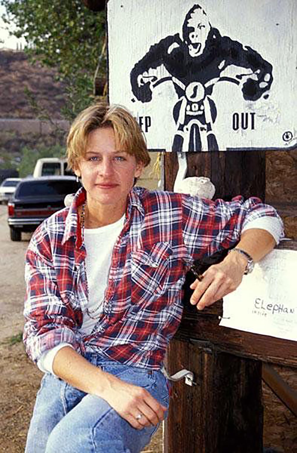 A person in a red plaid shirt and jeans sits casually outdoors, leaning against a wooden post. Behind them is a black and white sign with a gorilla on a motorcycle and the words “KEEP OUT.” A pickup truck is parked in the background.