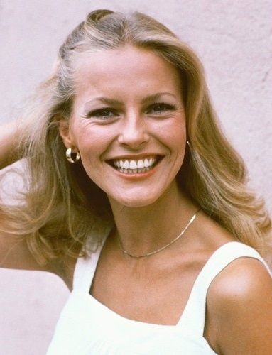 A smiling woman with long blonde hair wearing a white sleeveless top, gold hoop earrings, and a delicate necklace, stands against a pale pink background. She is touching her hair with one hand.