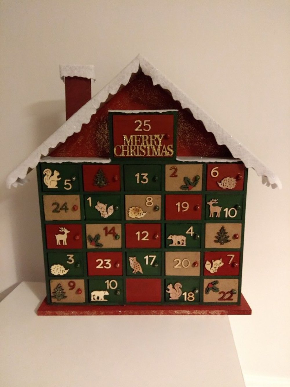 A wooden advent calendar shaped like a house with 25 numbered drawers decorated with animal and tree illustrations. The roof is dusted with white, and a red "Merry Christmas" sign is above the top drawer.