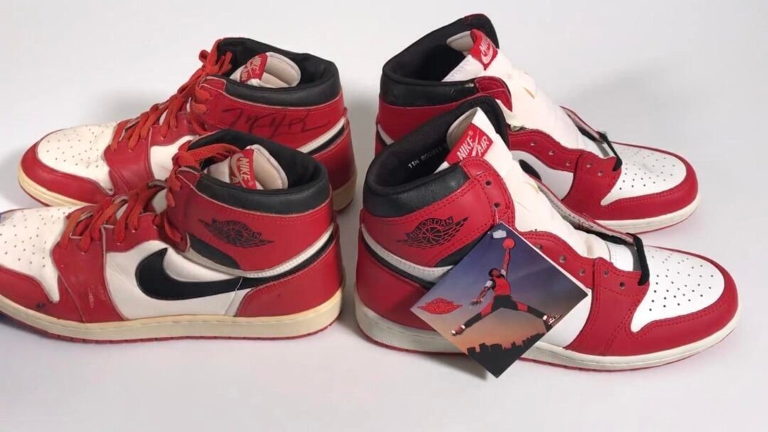 Two pairs of red, white, and black Nike Air Jordan 1 sneakers are displayed on a white surface. One pair looks worn and vintage; the other pair appears new with a tag featuring the Jumpman logo.