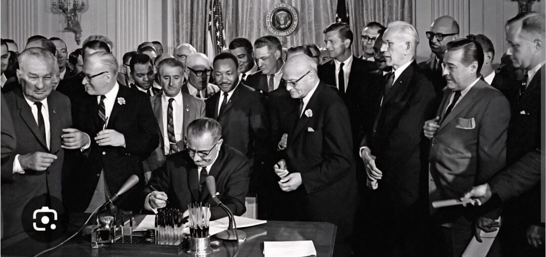 A group of men in suits stand around a seated man signing a document at a desk in an ornate room with American flags and microphones, suggesting a historic or official event.