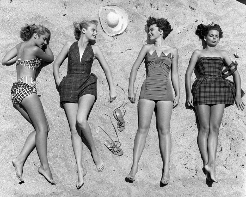 Four women in vintage swimsuits lie on the sand at the beach, looking relaxed and smiling at each other. A hat and sandals are placed between them. The image is in black and white.