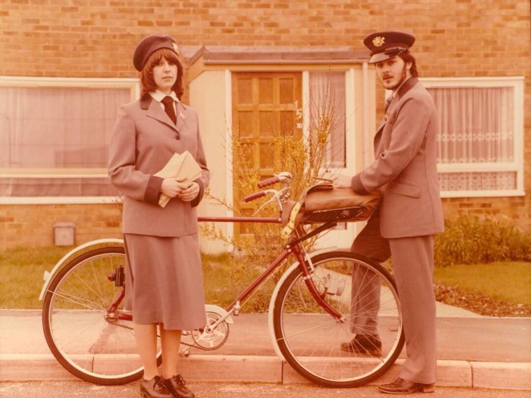 Two postal workers in vintage uniforms stand beside a bicycle in front of a house. One holds letters, and the other leans on the bicycle, both wearing hats and formal jackets. The scene appears to be from the past.