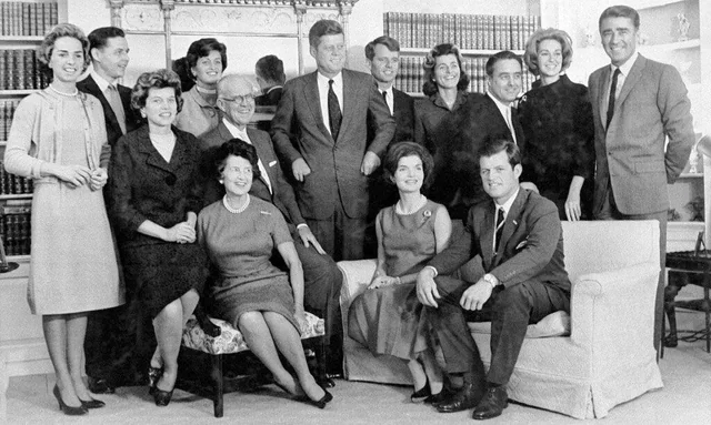A black-and-white photo of a large family group, with men and women dressed formally, seated and standing together in a room with bookshelves and a lamp in the background.